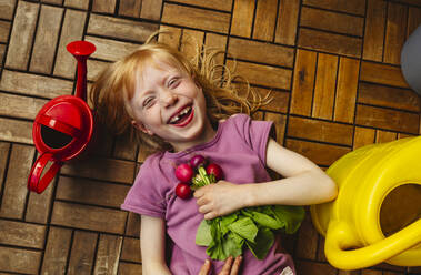 Cheerful girl with radish lying by watering can on balcony floor - IHF00840