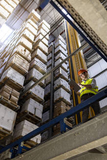 Smiling worker wearing hardhat using tablet PC standing by railing in warehouse - PESF03800