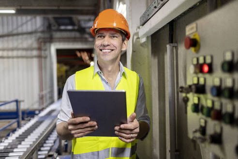 Happy worker wearing hardhat standing with tablet PC in warehouse - PESF03807