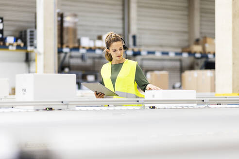 Young worker with tablet PC examining box on conveyor belt in warehouse - PESF03821