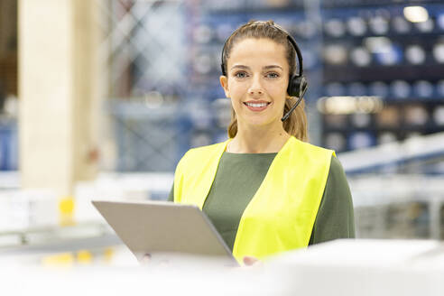 Smiling young worker wearing headset standing with tablet PC in warehouse - PESF03827