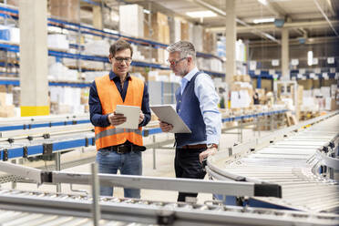 Worker discussing over tablet PC with manager standing by conveyor belt in warehouse - PESF03834