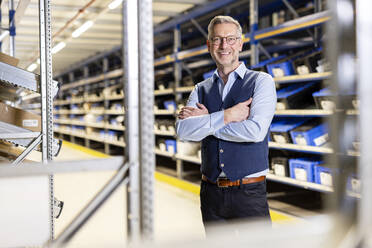 Happy manager wearing eyeglasses standing with arms crossed in warehouse - PESF03842