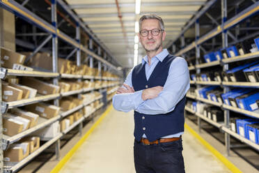 Smiling confident manager with arms crossed standing in aisle at warehouse - PESF03846