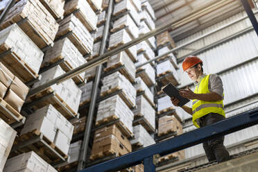 Mature worker wearing hardhat using tablet PC by railing in warehouse - PESF03880