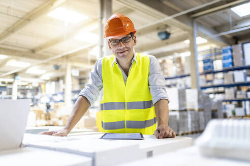 Smiling worker wearing eyeglasses leaning on box in warehouse - PESF03882