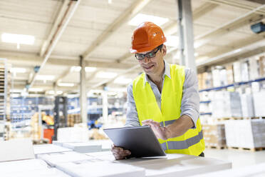 Lächelnder Arbeiter, der einen Tablet-PC benutzt und sich auf eine Kiste im Lager stützt - PESF03884