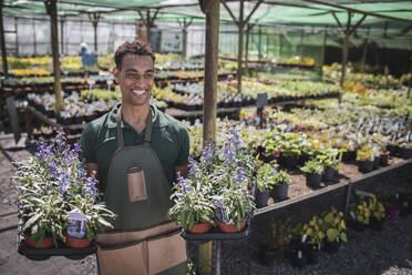 Glücklicher männlicher Gartenladenbesitzer mit Tabletts voller Topfblumen - CAIF32582