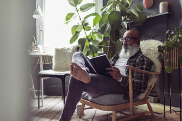 Barefoot mature man with beard reading book at home - CAIF32631