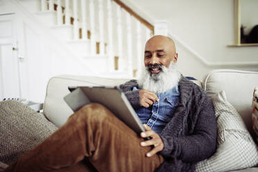 Mature man with beard using digital tablet on living room sofa - CAIF32646