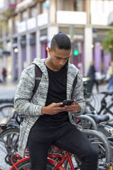Fokussierter hispanischer Mann in Freizeitkleidung, der eine SMS auf seinem Handy schreibt, während er auf einem Fahrrad auf der Straße einer Stadt mit einem Gebäude sitzt - ADSF34786