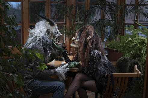 Krampus and witch with wineglass holding hands sitting at table - FCF02050