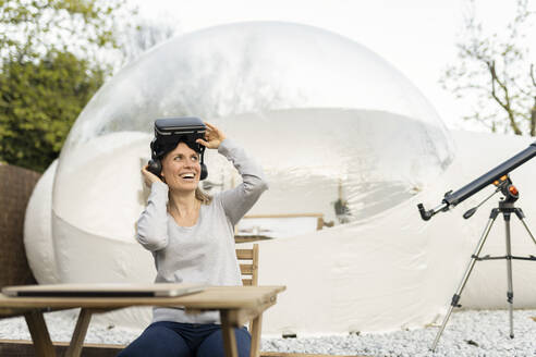 Glückliche Frau mit Virtual-Reality-Simulator vor einer transparenten Hotelkuppel - JCCMF06381