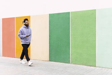 Man holding mobile phone listening music through headphones walking on footpath by colorful wall - PNAF03828