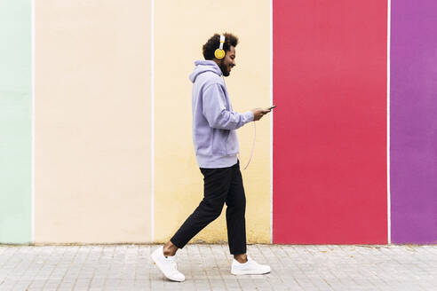 Lächelnder Mann, der ein Mobiltelefon benutzt und über Kopfhörer Musik hört, während er auf dem Fußweg geht - PNAF03829