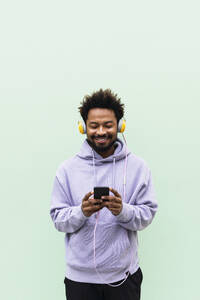 Smiling man using mobile phone and listening music through headphones against turquoise background - PNAF03830