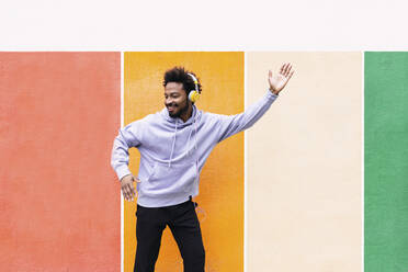 Happy man listening music through headphones and dancing in front of colorful wall - PNAF03840