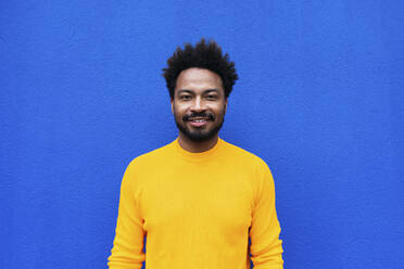 Smiling Afro man standing in front of blue wall - PNAF03849