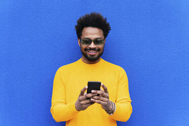 Lächelnder Afro-Mann, der ein Smartphone vor einer blauen Wand benutzt - PNAF03851