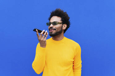 Smiling man talking on mobile phone through speaker in front of blue wall - PNAF03852