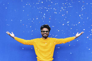 Smiling man with arms outstretched standing under falling confetti in front of blue wall - PNAF03853