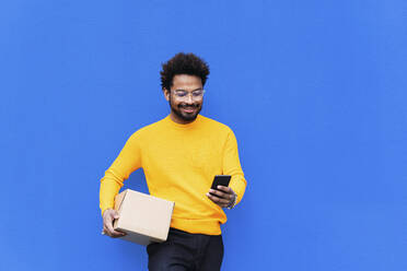 Lächelnder Zusteller, der eine Schachtel mit einem Smartphone vor einer blauen Wand hält - PNAF03854