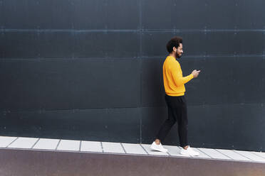 Lächelnder Mann, der ein Smartphone benutzt und auf einem Fußweg an einer schwarzen Wand spazieren geht - PNAF03870