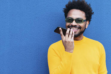 Smiling man talking on mobile phone through speaker in front of blue wall - PNAF03892