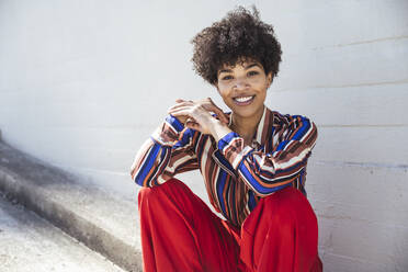 Lächelnde junge Frau mit Afrofrisur sitzt vor einer Wand - UUF26172