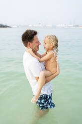 Smiling father carrying daughter standing in water at beach - SVKF00181