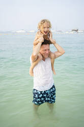 Father carrying daughter on shoulders standing in sea at beach - SVKF00182