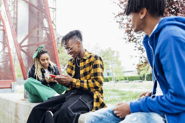 Glückliche multirassische Freunde mit Smartphone an der Wand sitzend - MEUF05647