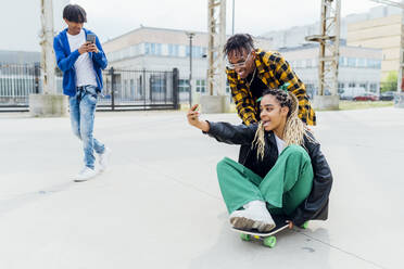 Junger Mann fotografiert Freund, der ein Selfie mit einer Frau macht, die auf einem Skateboard sitzt - MEUF05661