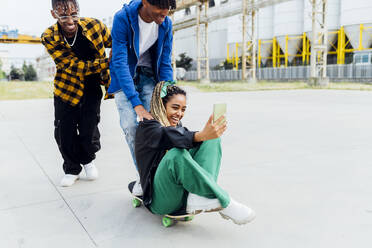 Fröhliche junge Frau, die auf einem Skateboard sitzt und ein Selfie mit Freunden auf einem Fußweg macht - MEUF05662