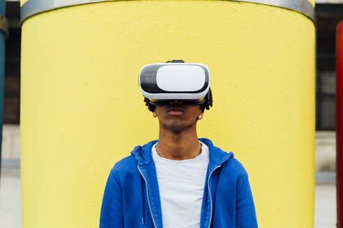 Junger Mann, der einen Virtual-Reality-Simulator trägt, steht vor einem gelben Betonrohr - MEUF05713