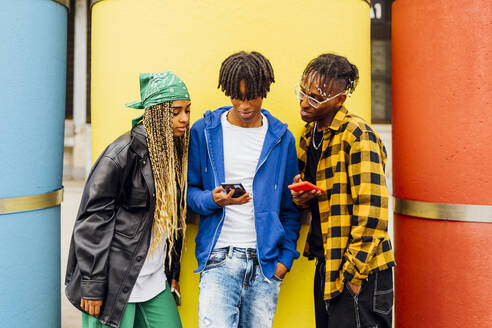 Junger Mann teilt Smartphone mit Freunden vor einem gelben Rohr - MEUF05723