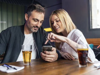 Smiling woman pointing at smart phone held by man sitting at table in restaurant - JSRF02060