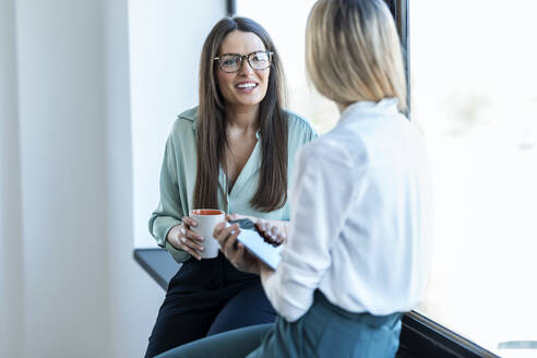 Lächelnde Geschäftsfrau mit Brille im Gespräch mit einem Kollegen, der im Büro am Fenster sitzt - JSRF02074