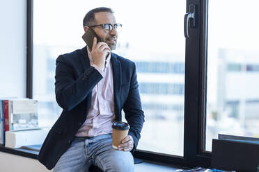 Geschäftsmann hält Einweg-Kaffeebecher und spricht mit seinem Smartphone, während er am Fenster im Büro sitzt - JSRF02080