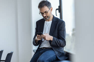 Businessman text messaging through smart phone sitting on window sill in office - JSRF02083