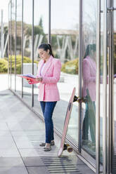 Geschäftsfrau, die vor einer Glaswand auf einem Skateboard stehend Dokumente liest - IFRF01663