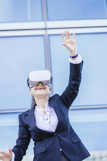 Lächelnde Geschäftsfrau mit Virtual-Reality-Simulator, die mit erhobener Hand vor einer Wand steht - IFRF01693