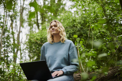 Contemplative freelancer sitting on fallen tree with laptop in forest - MMIF00317