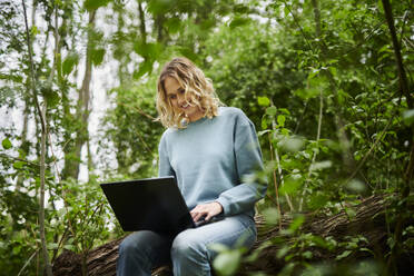 Lächelnder Freiberufler sitzt auf einem umgestürzten Baum und benutzt einen Laptop im Wald - MMIF00318