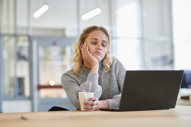 Müde junge Frau sitzt am Schreibtisch im Büro mit Laptop - MMIF00322