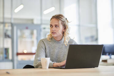 Junge Frau sitzt am Schreibtisch im Büro und benutzt einen Laptop - MMIF00323