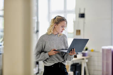 Junge Frau steht im Büro und benutzt einen Laptop - MMIF00324