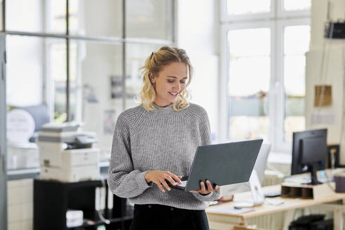 Lächelnde junge Frau, die im Büro steht und einen Laptop benutzt - MMIF00326