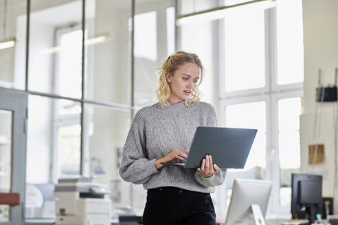 Young woman standing in office using laptop - MMIF00327