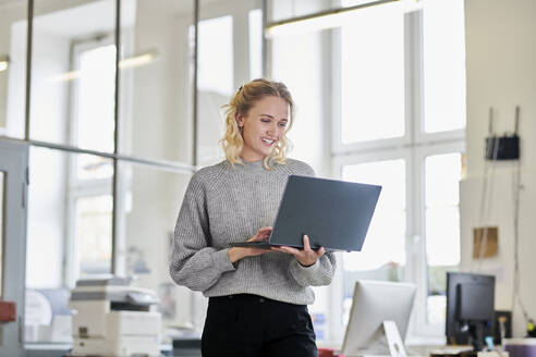 Happy young woman standing in office using laptop - MMIF00328